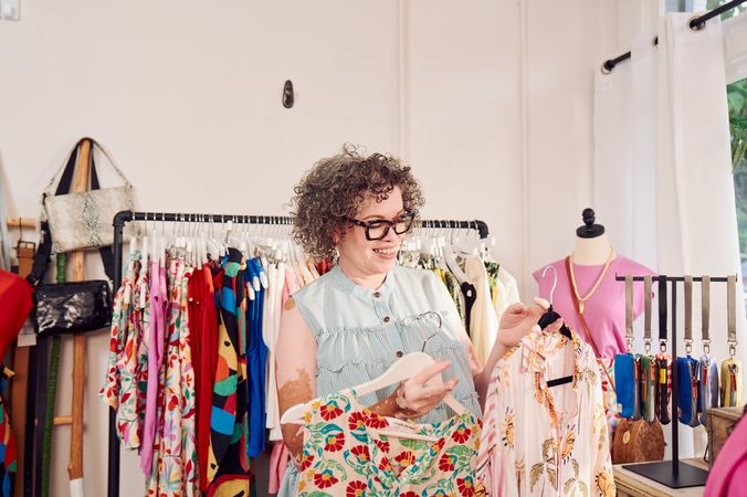 A Black woman shopkeeper with Vitiligo smiles and holds a patterned blouse in each hand, looking down at both options. She has a short, curly bob and is wearing glasses, a striped flowy blouse. Behind her are several clothing racks with high end garments, purses, belts, and mannequins.