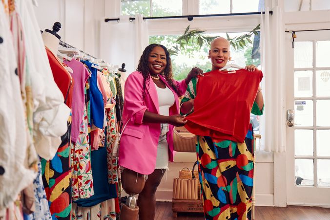 A bald African American person holds a black patterned skirt to her hips, deciding how it would look on her. Her friend, a black woman with a prosthetic leg, holds up a red shirt to her friend's torso, seeing how it would match the patterned skirt. The two friends are in a boutique. The black woman with a prosthetic leg has long, black curly hair and is wearing a vibrant pink blazer, and jean cut off shorts. The bald person is wearing a white and green striped crochet outfit and pointed white shoes. Clothing racks filled with high end garments are next to the women.