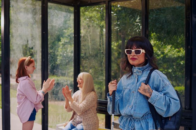 An Asian Filipino blind woman with Retinitis Pigmentosa and Usher Syndrome stands at a covered bus stop. She is wearing white sunglasses and is holding her white cane with her tattooed hands. She has on a jean jacket and a black backpack. Behind her, a white European girl with red hair plays pattycake hand game with her grandmother, who is sitting on the bus stop bench. Behind the bus stop are several large trees and walking paths.