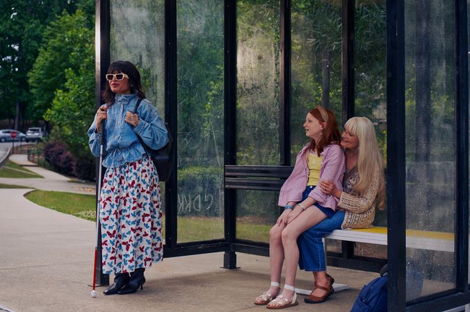 An Asian Filipino blind woman with Retinitis Pigmentosa and Usher Syndrome stands at a covered bus stop. She is wearing white sunglasses and is holding her white cane with her tattooed hands. She has on a jean jacket, a butterfly patterned skirt, and a black backpack. Behind her, a white European girl with red hair sits on her grandmother's lap on the bus stop bench. Behind the bus stop are several large trees and walking paths.