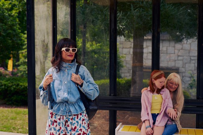 An Asian Filipino blind woman with Retinitis Pigmentosa and Usher Syndrome stands at a covered bus stop. She is wearing white sunglasses and is holding her white cane with her tattooed hands. She has on a jean jacket, a butterfly patterned skirt, and a black backpack. Behind her, a white European girl with red hair sits on her grandmother's lap on the bus stop bench and shows her a bracelet she is wearing. Behind the bus stop are several large trees, a landscaped garden, and a stone building.