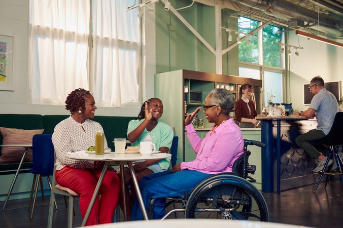An African American daughter and auntie give each other a high five at a table in a modern cafe, while the girl's mother smiles and watches. The auntie is paraplegic and is seated in a manual wheelchair.  She has short gray hair and is wearing a lavender silk blouse and dark blue jeans. The mother has short tightly curled red hair and is wearing a patterned blouse and red pants. The daughter is wearing a cochlear implant and has her braided hair pulled back into a pony tail. She is wearing a light green cardigan and jean shorts. Behind them, a barista prepares drinks and a man types on a laptop.