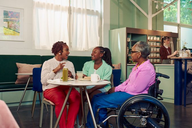 An African American mother and daughter chat at a table in a modern cafe, while the girl's auntie smiles and watches. The auntie is paraplegic and is seated in a manual wheelchair. She has short gray hair and is wearing a lavender silk blouse and dark blue jeans. The mother has short tightly curled red hair and is wearing a patterned blouse and red pants. The daughter is wearing a cochlear implant and has her braided hair pulled back into a pony tail. She is wearing a light green cardigan and jean shorts. Behind them, a barista prepares drinks.
