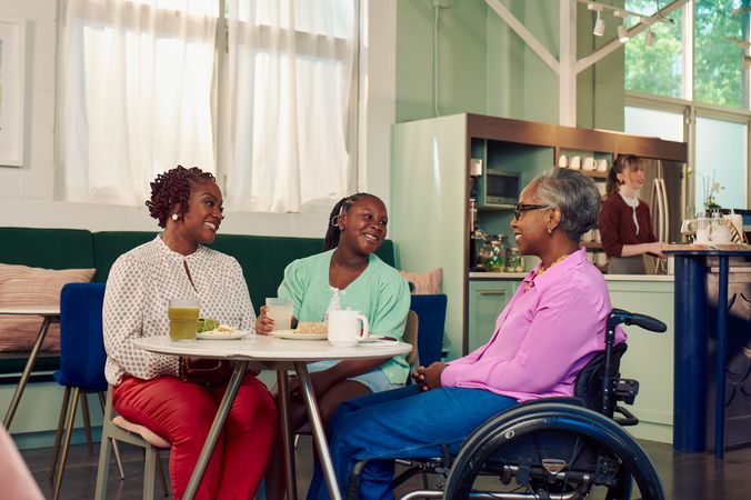 An African American mother, daughter, and auntie are smiling while chatting at a table in a modern cafe. The auntie is paraplegic and is seated in a manual wheelchair. She has short gray hair is wearing a lavender silk blouse and dark blue jeans. The mother has short tightly curled red hair and is wearing a patterned blouse and red pants. The daughter is wearing a cochlear implant and has her braided hair pulled back into a pony tail. She is wearing a light green cardigan and jean shorts. Behind them, a barista prepares drinks and a man types on a laptop.