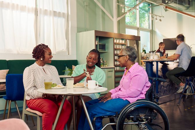 An African American girl shows her auntie something on her phone at a table in a modern cafe, while the girl's mother smiles broadly and watches. The auntie is paraplegic and is seated in a manual wheelchair. She has short gray hair is wearing a lavender silk blouse and dark blue jeans. The mother has short tightly curled red hair and is wearing a patterned blouse and red pants. The daughter is wearing a cochlear implant and has her braided hair pulled back into a pony tail. She is wearing a light green cardigan and jean shorts. Behind them, a barista hands a drink to a man seated the cafe bar.