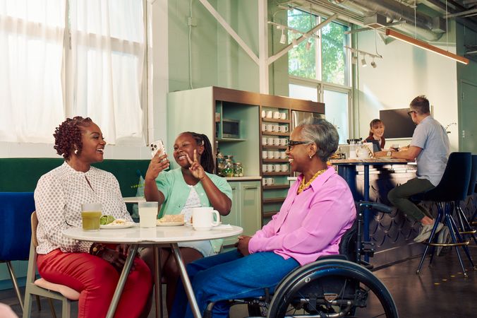An African American girl takes a selfie and holds up two fingers in a peace sign in a modern cafe, while the girl's motherand auntie watch. The auntie is paraplegic and is seated in a manual wheelchair. She has short gray hair is wearing a lavender silk blouse and dark blue jeans. The mother has short tightly curled red hair and is wearing a patterned blouse and red pants. The daughter is wearing a cochlear implant and has her braided hair pulled back into a pony tail. She is wearing a light green cardigan and jean shorts. Behind them, a barista hands a drink to a man seated the cafe bar.