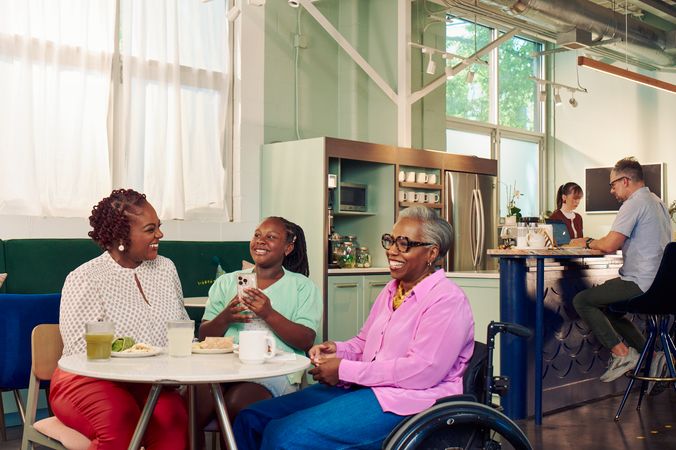 An African American girl shows smiles and holds her phone at a table in a modern cafe, while the girl's mother and auntie laugh together. The auntie is paraplegic and is seated in a manual wheelchair. She has short gray hair is wearing a lavender silk blouse and dark blue jeans. The mother has short tightly curled red hair and is wearing a patterned blouse and red pants. The daughter is wearing a cochlear implant and has her braided hair pulled back into a pony tail. She is wearing a light green cardigan and jean shorts. Behind them, a barista makes drinks and a man types on a laptop.