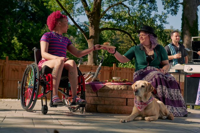 At a backyard barbeque, a white nonbinary person sits on a low stone table and passes a plate of food to a Black nonbinary person seated in a manual wheelchair with rainbow spokes. The white nonbinary person has a uniquely shapped dark gray rollator decorated with purple acrylic chains nearby. They have layered purple hair with two thin long braids on each side and are wearing a black hat, a green shirt, and a purple patterned skirt. The Black nonbinary person seated in their manual wheelchair has a curly pink fro with loc bangs and is wearing a purple and pink striped crop top, pink shorts, and purple sandals. They have several tattoos on both legs. A golden labrador retriever wearing a pink hankerchief sits at their feet. Behind them, another friend, a multi-ethnic man with curly black hair, holds a drink and chats near a grill.