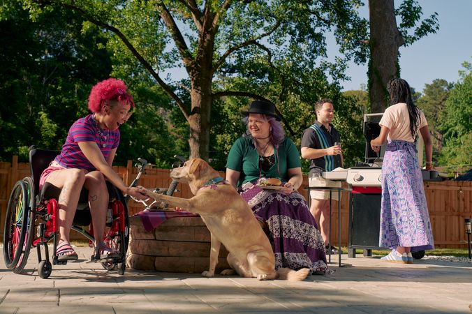At a backyard barbeque, a golden labrador retriever places her paw in the palm of a Black nonbinary person's hand. The Black nonbinary person is seated in a manual wheelchair with rainbow spokes and has their mouth open in praise of the dog. Next to them, a white nonbinary person sits on a low stone table and holds a plate of food. They have layered purple hair with two thin long braids on each side and are wearing a black hat, a green shirt, and a purple patterned skirt. The Black nonbinary person seated in their manual wheelchair has a curly pink fro with loc bangs and is wearing a purple and pink striped crop top, pink shorts, and purple sandals. They have several tattoos on both legs. Behind them, two friends chat at a grill. One friend is an African American woman and is holding a spatula. The other friend is a multi-ethnic man with curly black hair.