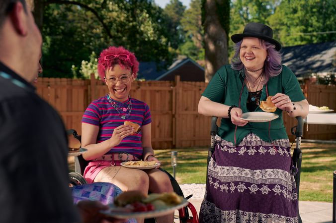At a backyard barbeque, two nonbinary people sit in their mobility aids, laughing and holding plates of food. One nonbinary person, a Black person with a curly pink fro with loc bangs wearing glasses is seated in a manual wheelchair. They are wearing bright pink sunglassese on their head, a purple and pink striped crop top, pink shorts, and purple sandals. They have several tattoos on both legs. The second nonbinary person, a white person with layered purple hair with two long thin red braids on each side, is seated on a dark gray rollator. They are wearing a black hat, a green shirt, and a purple patterned skirt.