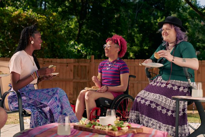 At a backyard barbeque, two nonbinary people sit in their mobility aids, chat with a friend while holding plates of food.  One nonbinary person, a Black person with a curly pink fro with loc bangs wearing glasses is seated in a manual wheelchair. They are wearing bright pink sunglassese on their head, a purple and pink striped crop top, pink shorts, and purple sandals. They have several tattoos on both legs. The second nonbinary person, a white person with layered purple hair with two long thin red braids on each side, is seated on a dark gray rollator. They are wearing a black hat, a green shirt, and a purple patterned skirt. Their friend, an African American woman with long braided hair, is wearing a pink shirt and patterned blue and purple skirt.