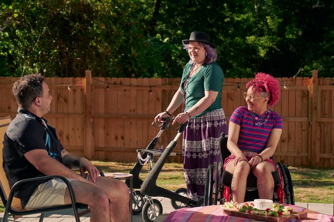 A white nonbinary person stands with their uniquely shaped dark gray rollator that is decorated with purple acrylic chains. They have purple layered hair with two long thin red braids on both sides and are wearing a black hat, green shirt, and purple patterned skirt. They chat with a friend, who is a multi-ethnic man with curly black hair, who is sitting in a patio chair. Next to them is a Black nonbinary person seated in their manual wheelchair with rainbow spokes. They have a curly pink fro with loc bangs and are wearing bright pink sunglassese on their head, a purple and pink striped crop top, pink shorts, and purple sandals. They have several tattoos on both legs.