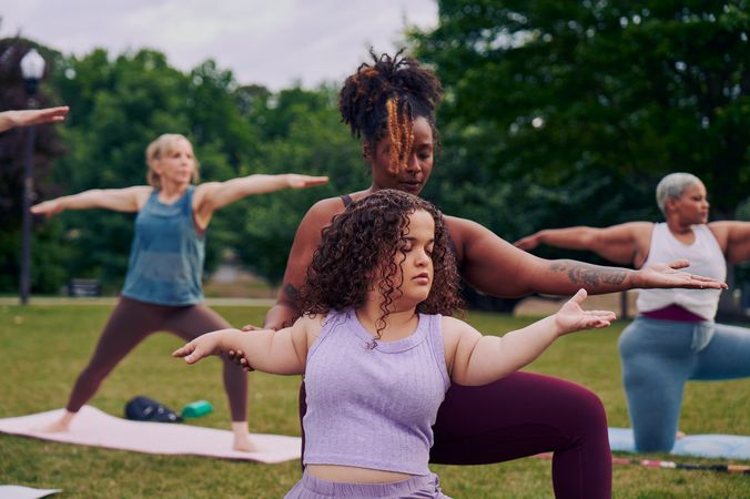 At an outdoor yoga class, a young white woman with dwarfism stands in Warrior pose, with her eyes closed, both arms extended out parallel to the ground, one facing forward and one reaching back. A Black woman yoga instructor kneels behind her and gently guides her arms into the pose. The student has long, curly brown hair and is wearing a purple ribbed tank with comfortable, loose purple pants. The instructor has her black hair pulled back into a natural ponytail with dyed bangs and is wearing a black tank and maroon yoga pants. Behind them, two tall women sit on their mats in the same pose. One of the women in the background is in a modified form of the pose, kneeling. The class is in a grassy park with many large trees.
