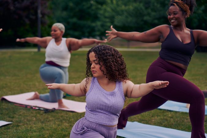 At an outdoor yoga class, a young white woman with dwarfism stands in Warrior pose, with her eyes closed, both arms extended out parallel to the ground, one facing forward and one reaching back. She has long, curly brown hair and is wearing a purple ribbed tank with comfortable, loose purple pants. Behind her, a tall Black woman instructor smiles while also in the same pose. Another Black woman is in a modified form of the pose, kneeling. Her cane is next to her mat. The class is in a grassy park with many large trees.