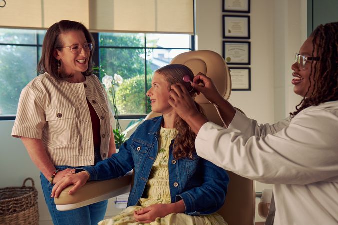 A young white deaf teen with braces sits in a medical treatment chair while an audiologist places a mauve colored cochlear implant on her head. The teen looks at her mother, who is standing next to her in the office. The teen has long, wavy sandy blonde hair and is wearing a jean jacket, and a yellow blouse and skirt. The audiologist is a Black deafBlind woman wearing glasses and has dark brown shoulder length locs. The teen's mother is a middle-aged white woman with straight brown hair and glasses. She is wearing an off-white striped summer jacket and jeans. A row of four framed professional degrees are hanging on the wall behind them.