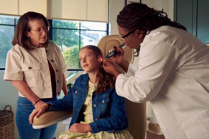 A Black deafBlind audiologist with a cochlear implant and glasses smiles and uses an otoscope to look into the ears of a young white deaf teen with braces seated in a medical treatment chair. The teen is softly smiling and has long, wavy sandy blonde hair and is wearing a jean jacket, and a yellow blouse. The teen's mother is standing next to her, observing the audiologist use the tool.