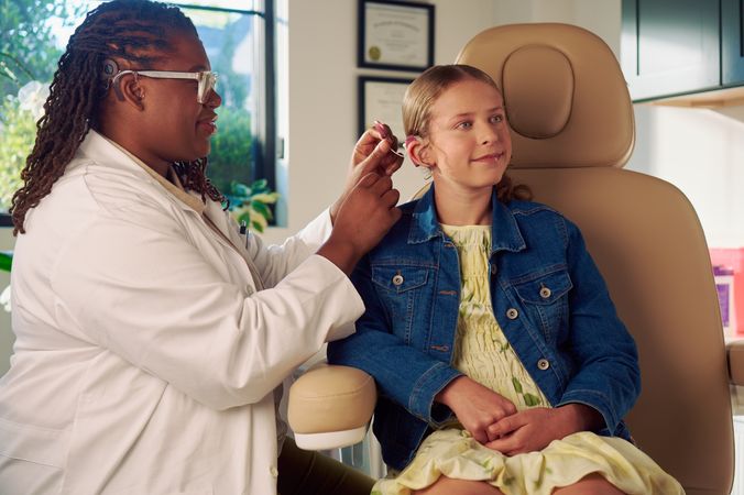 A Black deafBlind audiologist with a cochlear implant and glasses places a cochlear implant on a young white deaf teen with braces seated in a medical treatment chair. The teen is smiling and has long, wavy sandy blonde hair and is wearing a jean jacket, and a yellow blouse and skirt. Framed professional degrees are hanging on the wall behind them.