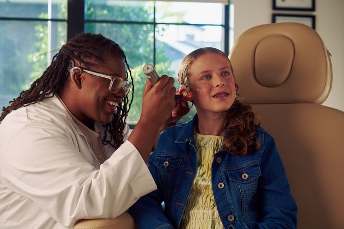 A Black deafBlind audiologist with a cochlear implant and glasses smiles and uses an otoscope to look into the ears of a young white deaf teen with braces seated in a medical treatment chair. The teen is smiling and has long, wavy sandy blonde hair and is wearing a jean jacket, and a yellow blouse and skirt. Framed professional degrees are hanging on the wall behind them.