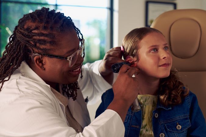 A Black deafBlind audiologist with a cochlear implant and glasses smiles and uses an otoscope to look into the ears of a young white deaf teen with braces seated in a medical treatment chair. The teen is softly smiling and has long, wavy sandy blonde hair and is wearing a jean jacket, and a yellow blouse.
