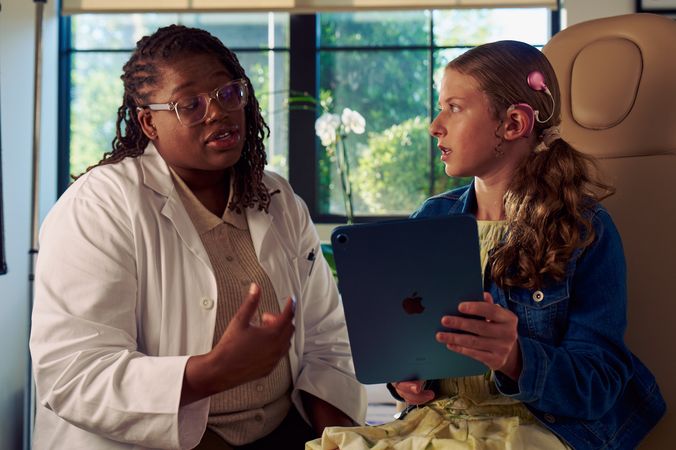A Black deafBlind audiologist with a cochlear implant and glasses talks with a young white deaf teen with braces holding a tablet. The teen is seated in a medical treatment chair and looks at the audiologist with curiosity. She and has long, wavy sandy blonde hair and is wearing a jean jacket, and a yellow blouse and skirt.
