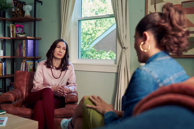 An Eastern European white woman therapist sits in a large chair and looks at a client seated on a couch in front of her. The client is a Latina woman with dark brown wavy hair pulled into a ponytail. The therapist's office has several books on display, an abstract art piece, and a window letting in natural light. The therapist is wearing a silk pink top, maroon dress pants, and a minimalist necklace. The client is wearing gold hoop earrings, a jean jacket, green skirt, and sneakers.