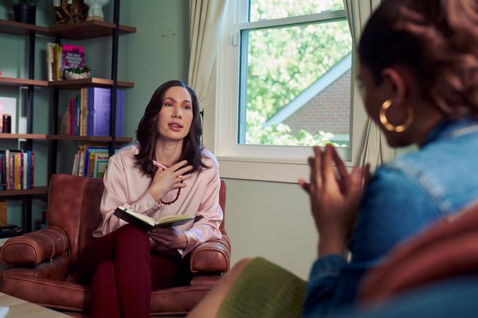An Eastern European white woman therapist sits in a large chair and looks at a client seated on a couch in front of her. She is holding a pen and open notebook. The client is a Latina woman with dark brown wavy hair pulled into a ponytail. The therapist's office has several books on display and a window letting in natural light. The therapist is wearing a silk pink top, maroon dress pants, and a minimalist necklace. The client is wearing gold hoop earrings, a jean jacket, and green skirt.