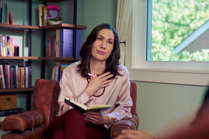 An Eastern European white woman therapist sits in a large chair and holds her hand up to her chest with an understanding look on her face. She is holding a pen and open notebook, and is looking at a client off camera. The therapist's office has several books on display and a window letting in natural light. The therapist is wearing a silk pink top, maroon dress pants, and a minimalist necklace.