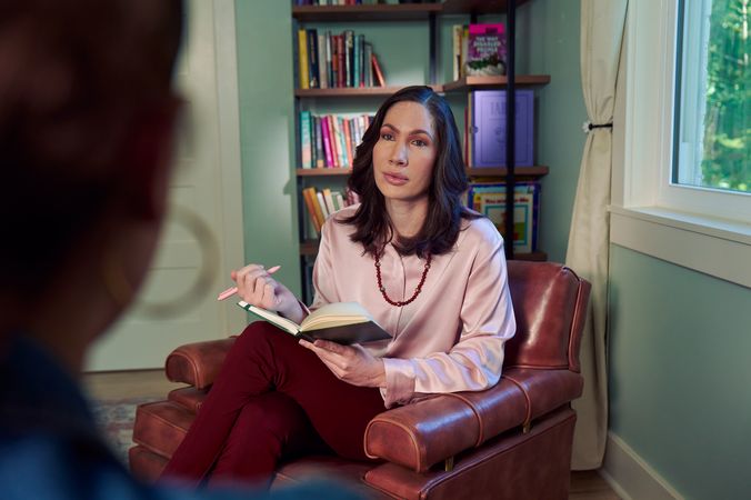 An Eastern European white woman therapist sits in a large chair with an understanding look on her face. She is holding a pen and open notebook, and is looking at a client off camera. The therapist's office has several books on display and a window letting in natural light. The therapist is wearing a silk pink top, maroon dress pants, and a minimalist necklace.