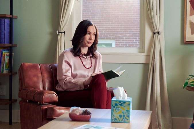 An Eastern European white woman therapist sits in a large chair looks at a client off camera. She is holding an open notebook. A tissue box and a red marble heasrt-shaped bowl of crystals are on a low coffee table in front of her. The therapist's office has several books on display and a window letting in natural light. The therapist is wearing a silk pink top, maroon dress pants, and a minimalist necklace.