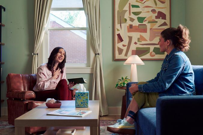 An Eastern European white woman therapist sits in a large chair and looks at a client seated on a couch in front of her. She is holding her hand to her chest and smiles at her client knowingly. The client is a Latina woman with dark brown wavy hair pulled into a ponytail. A tissue box, a red marble heasrt-shaped bowl of crystals, and a magazine are on a low coffee table in the center of the office. An abstract art piece, a lamp, and a small house plant are next and a window letting in natural light. The therapist is wearing a silk pink top, maroon dress pants, and a minimalist necklace. The client is wearing gold hoop earrings, a jean jacket, green skirt, and sneakers.