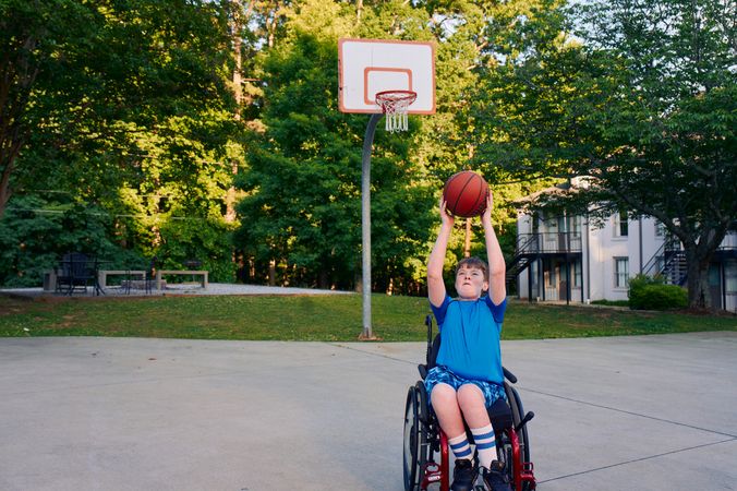 A white teen boy seated in a manual wheelchair holds a basketball over his head, with a look of concentration on his face. He is playing at a basketball court at a neighborhood park. He has short brown hair and is wearing blue athletic wear.