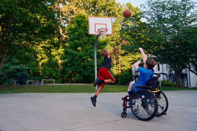 Two teens play basketball at a neighborhood park. One teen, a white boy seated in a manual wheelchair, shoots the ball. The other teen, a light-skinned African American male, jumps forward with arms outstretched, defending the shot. The teen seated in a manual wheelchair has short brown hair and is wearing blue athletic wear. The other teen is wearing black and red athletic wear.