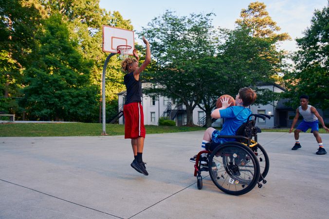 Three teens play basketball at a neighborhood park. One teen, a white boy seated in a manual wheelchair, holds the ball, ready to shoot. A second teen, a light-skinned African American male, jumps forward with arms outstretched, ready to defend the shot. A third teen, a Black boy, crouches, ready to receive a pass. The teen seated in a manual wheelchair has short brown hair and is wearing blue athletic wear. The second teen defending has curly brown to blonde hair is wearing black and red athletic wear. The third teen has short natural black hair styled high on top and is wearing gray and blue athletic wear.