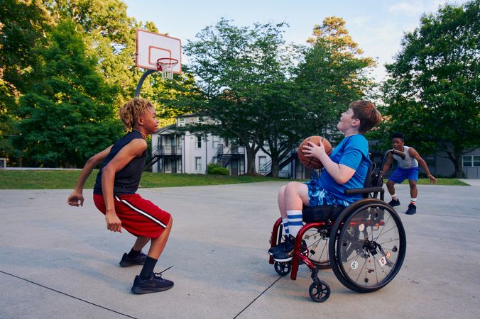Three teens play basketball at a neighborhood park. One teen, a white boy seated in a manual wheelchair, holds the ball, ready to shoot. A second teen, a light-skinned African American male, crouches in a jumping position. A third teen, a Black boy, crouches, ready to receive a pass. The teen seated in a manual wheelchair has short brown hair and is wearing blue athletic wear. The second teen ready to jump has curly brown to blonde hair is wearing black and red athletic wear. The third teen has short natural black hair styled high on top and is wearing gray and blue athletic wear.