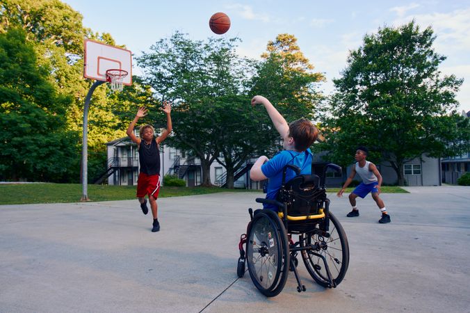 Two teens play basketball at a neighborhood park. One teen, a white boy seated in a manual wheelchair, shoots the ball. The other teen, a light-skinned African American male, jumps forward with arms outstretched, defending the shot. The teen seated in a manual wheelchair has short brown hair and is wearing blue athletic wear. The other teen is wearing black and red athletic wear.