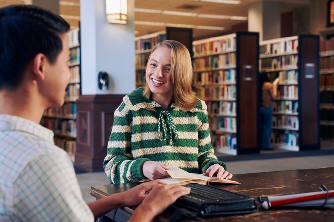 A white teenage girl seated in a manual wheelchair smiles brightly at a friend and holds a book open at a table in a library. Her friend, a Chinese teen, who is a blind cane user, has a refreshable braille display in front of him. His white cane is collapsed and is placed the table next to him. The teen girl has a dark blonde bob and is wearing a green and white striped knit shirt and jeans. The teen boy has slicked back black hair and is wearing a white plaid button down shirt.