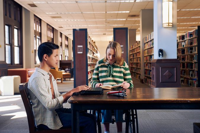 Two teenage friends read at a table in a library. One teen, a Chinese teen, who is a blind cane user, reads on a refreshable braille display. His white cane is collapsed and is placed the table next to him. The other teen, a white teenage girl seated in a manual wheelchair, is seated in a manual wheelchair and holds a hardcover book open. The teen girl has a dark blonde bob and is wearing a green and white striped knit shirt and jeans. The teen boy has slicked back black hair and is wearing a white plaid button down shirt.
