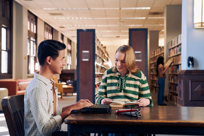 Two teenage friends laugh and read at a table in a library. One teen, a Chinese teen, who is a blind cane user, reads on a refreshable braille display. His white cane is collapsed and is placed the table next to him. The other teen, a white teenage girl seated in a manual wheelchair, is seated in a manual wheelchair and looks down at a hardcover book she has open. The teen girl has a dark blonde bob and is wearing a green and white striped knit shirt and jeans. The teen boy has slicked back black hair and is wearing a white plaid button down shirt.