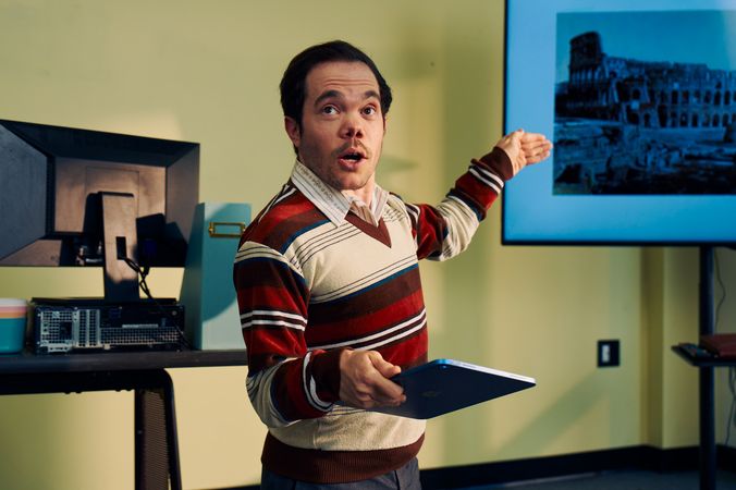 A white male professor with medium brown hair and light brown eyes who has dwarfism gestures at a large monitor next to him, displaying a black and white photo of the Colosseum. He is holding a tablet in his other hand. He is dressed in a striped V-neck sweater, charcoal dress pants, and dressy-casual shoes, standing with a warm, confident presence. Behind him, his desk has a computer, a file holder, and a mug.