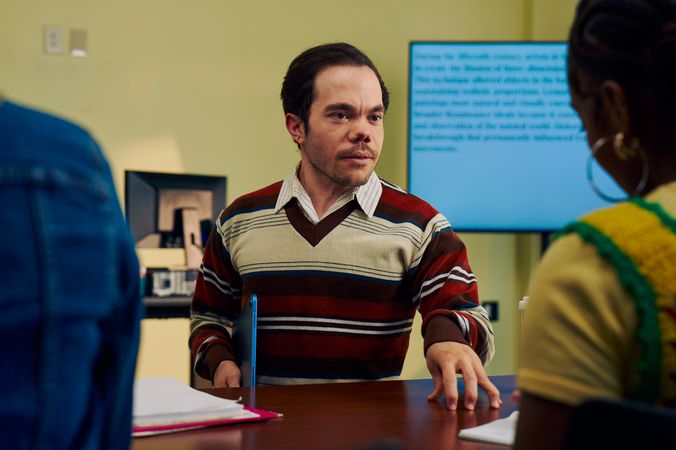 A white male professor with medium brown hair and light brown eyes who has dwarfism stands and talks to a student with focus. He is holding a tablet with one hand and taps the student's desk with the other. The student takes notes as the professor speaks. The professor is wearing a striped V-neck sweater, charcoal dress pants, and dressy-casual shoes. The student is a Black woman with hair pulled into a bun and is wearing a yellow shirt. Behind them is a large monitor with text displayed.