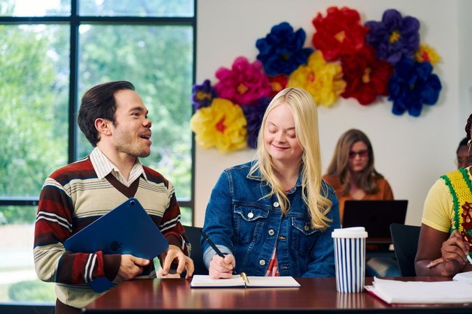 A caucasian college student with Down Syndrome sits at a lecture table and takes notes while her professor, who is standing next to her, chats with her. She has long blonde hair, blue eyes, and is wearing a jean hacket and several bright scrunchies on her wrist. Another student behind her, a blind white woman wearing glasses with red lenses, types on a laptop. A large flower sculpture hangs on the wall behind the desks. Her professor, a white man with medium brown hair and light brown eyes who has dwarfism, smiles and holds a tablet in his hand. He has short brown hair and is wearing a red striped sweater, a collared shirt, and dark dress pants.
