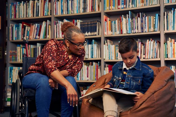 An African American teacher seated in a manual wheelchair smiles and watches a young student sitting in a bean bag chair read a picture book. The teacher has medium brown skin, reddish brown hair pulled into a short natural ponytail, and is wearing reading glasses, a red patterned blouse, and jeans. The young student is a Hispanic and Mediterranean-American boy with short brown hair. He  has olive skin and is wearing a blue striped shirt, a jean jacket, and khaki pants. A wall of children's books are behind them.