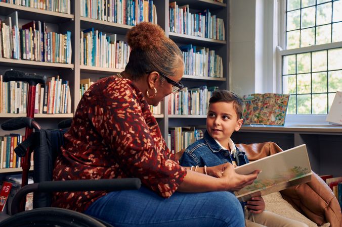 A young boy sitting in a bean bag chair looks up at his teacher, an African American woman seated in a manual wheelchair. They are both holding a picture book. The teacher has a red walking cane fastened to the back of her wheelchair. She has medium brown skin, reddish brown hair pulled into a short natural ponytail, and is wearing reading glasses, a red patterned blouse, and jeans. The young student is a Hispanic and Mediterranean-American boy with short brown hair. He  has olive skin and is wearing a blue striped shirt, a jean jacket, and khaki pants. A wall of children's books are behind them.